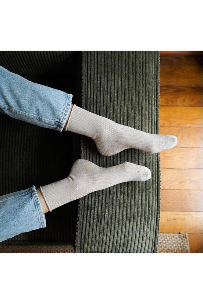 Person wearing beige glitter socks from Klue and blue jeans sitting on a dark green cushioned chair.
