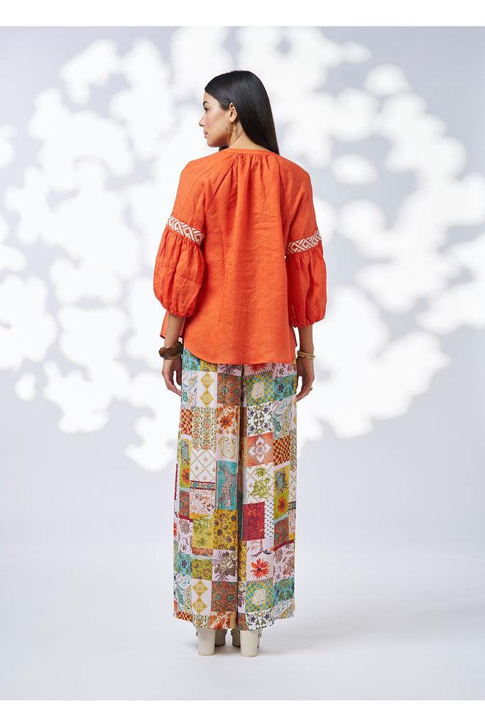 Woman wearing an orange top and colourful patchwork pants against a white background. Loobies Story Maya Top Burnt Orange.