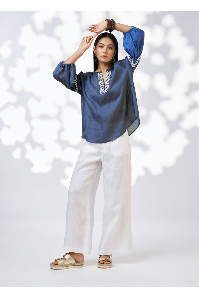 Woman wearing an blue denim top and white pants against a white background. Loobies Story Maya Top Denim.