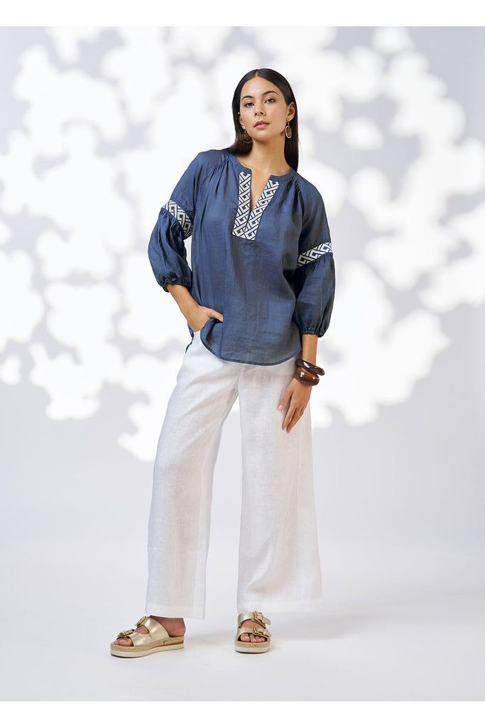 Woman wearing an blue denim top and white pants against a white background. Loobies Story Maya Top Denim.
