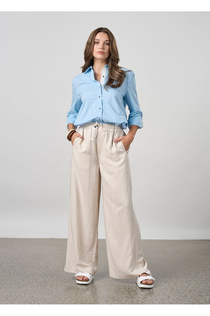 Woman wearing a light blue shirt and beige pants against a plain background. Madly Sweetly Eclipse Pant in Chalk.