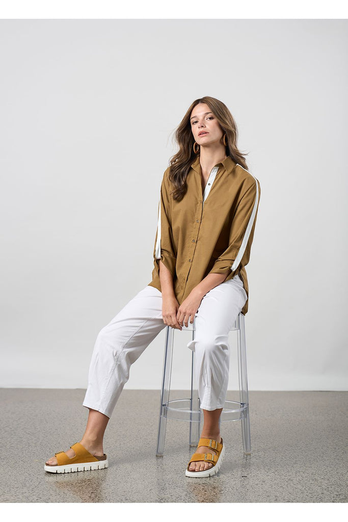 Woman wearing a brown shirt and white pants standing against a plain background. Madly Sweetly Ratio Pant in White.