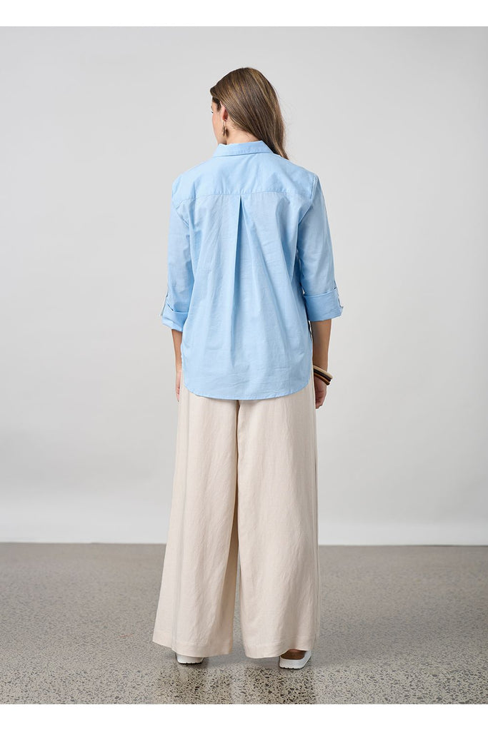 Woman wearing a light blue shirt and beige pants against a plain background. Madly Sweetly Eclipse Pant in Chalk.