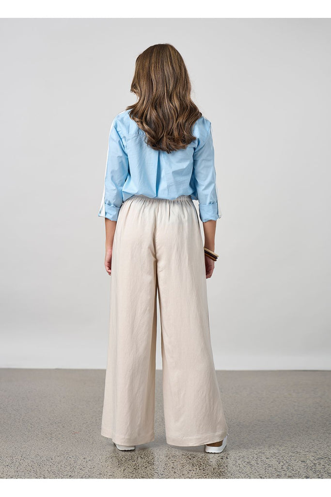 Woman wearing a light blue shirt and beige pants against a plain background. Madly Sweetly Eclipse Pant in Chalk.