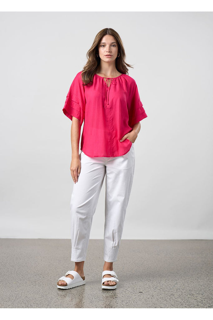 Woman wearing a pink blouse and white pants standing against a plain background. Madly Sweetly Ratio Pant in White.