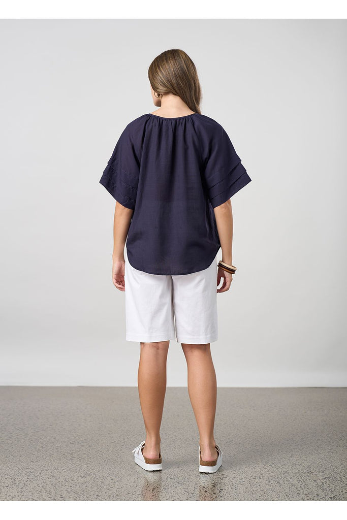 Woman wearing a navy blue blouse and white shorts on a light grey background. Madly Sweetly Parallel Top in Navy.