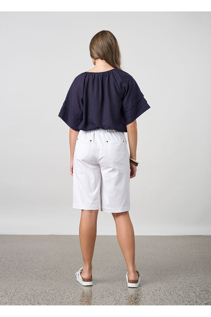 Woman wearing a navy blue blouse and white shorts on a light grey background. Madly Sweetly Parallel Top in Navy.