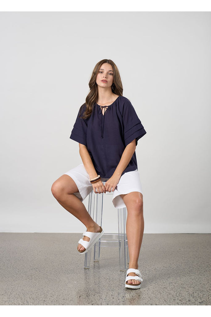 Woman wearing a navy blue blouse and white shorts on a light grey background. Madly Sweetly Parallel Top in Navy.