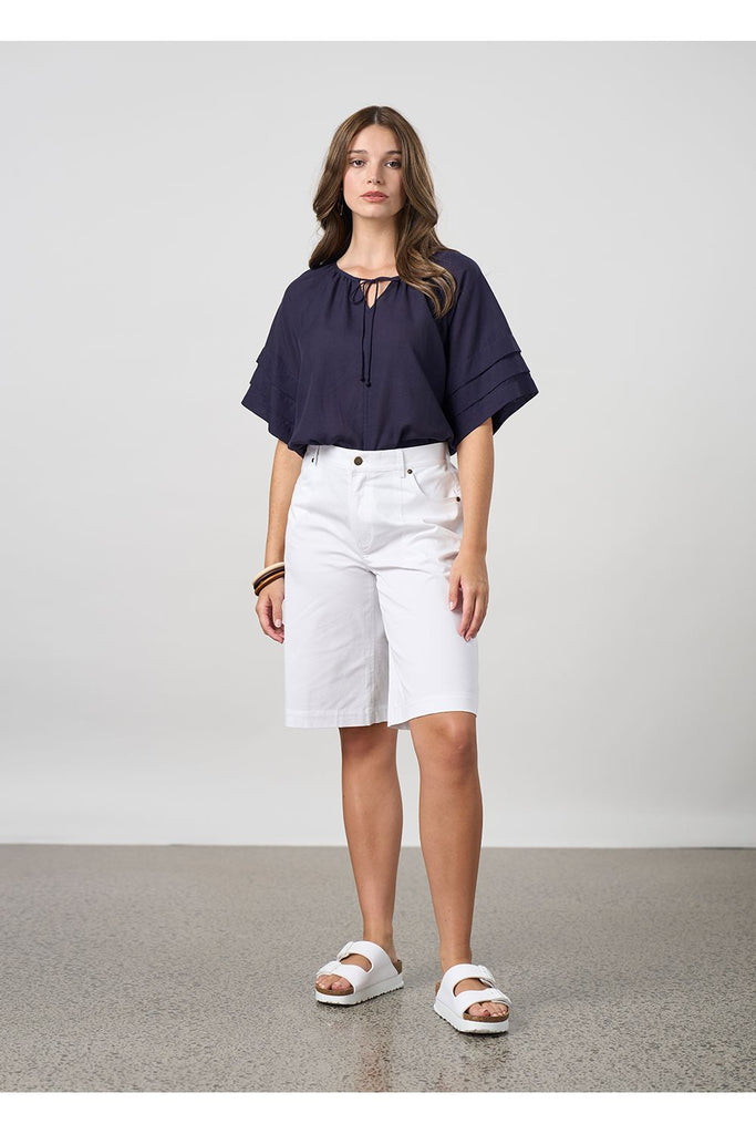 Woman wearing a navy blue blouse and white shorts on a light grey background. Madly Sweetly Parallel Top in Navy.