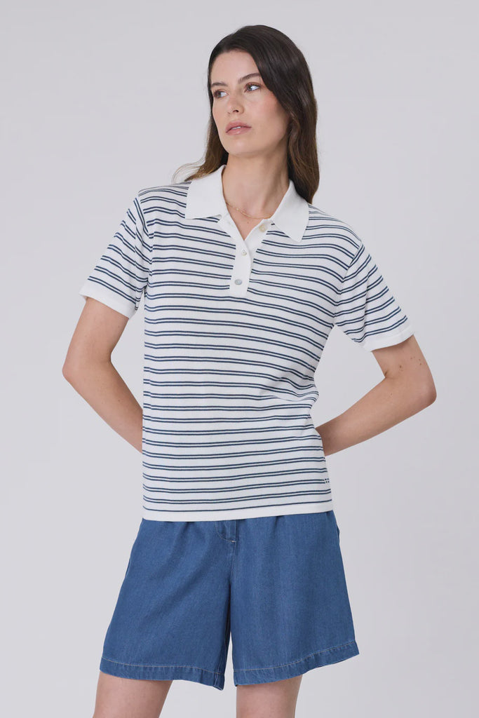 Woman wearing a striped polo shirt and blue shorts on a plain background. Marlow Chime Knit Polo Shirt in indigo Stripe.