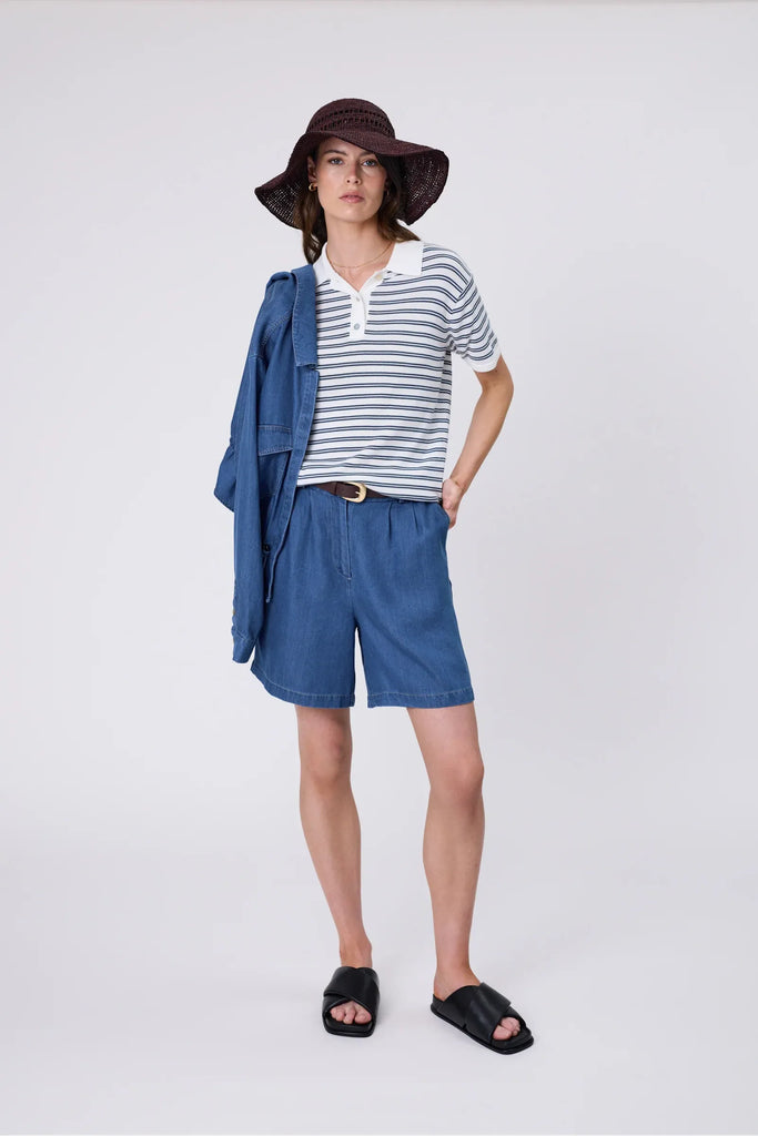 Woman wearing a striped polo shirt and blue shorts on a plain background. Marlow Chime Knit Polo Shirt in indigo Stripe.