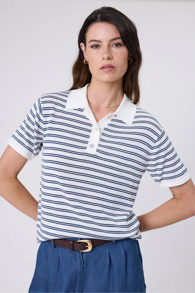 Woman wearing a striped polo shirt and blue shorts on a plain background. Marlow Chime Knit Polo Shirt in indigo Stripe.