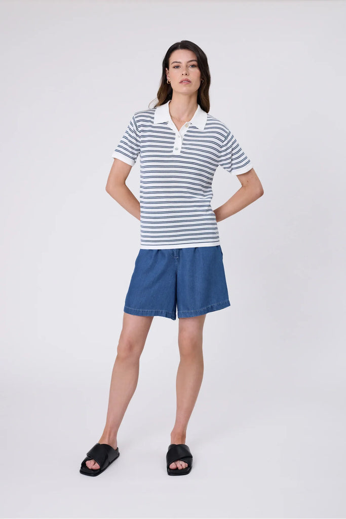 Woman wearing a striped polo shirt and blue shorts on a plain background. Marlow Chime Knit Polo Shirt in indigo Stripe.