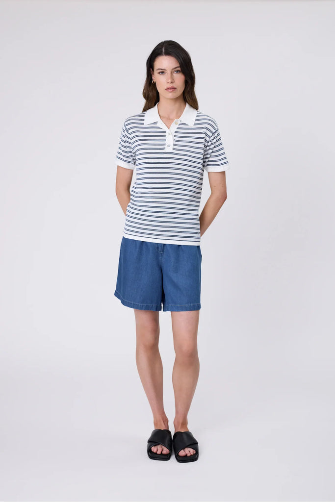 Woman wearing a striped polo shirt and blue shorts on a plain background. Marlow Chime Knit Polo Shirt in indigo Stripe.