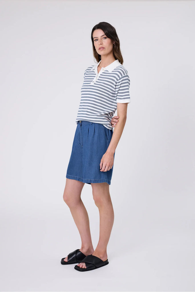 Woman wearing a striped polo shirt and blue shorts on a plain background. Marlow Chime Knit Polo Shirt in indigo Stripe.
