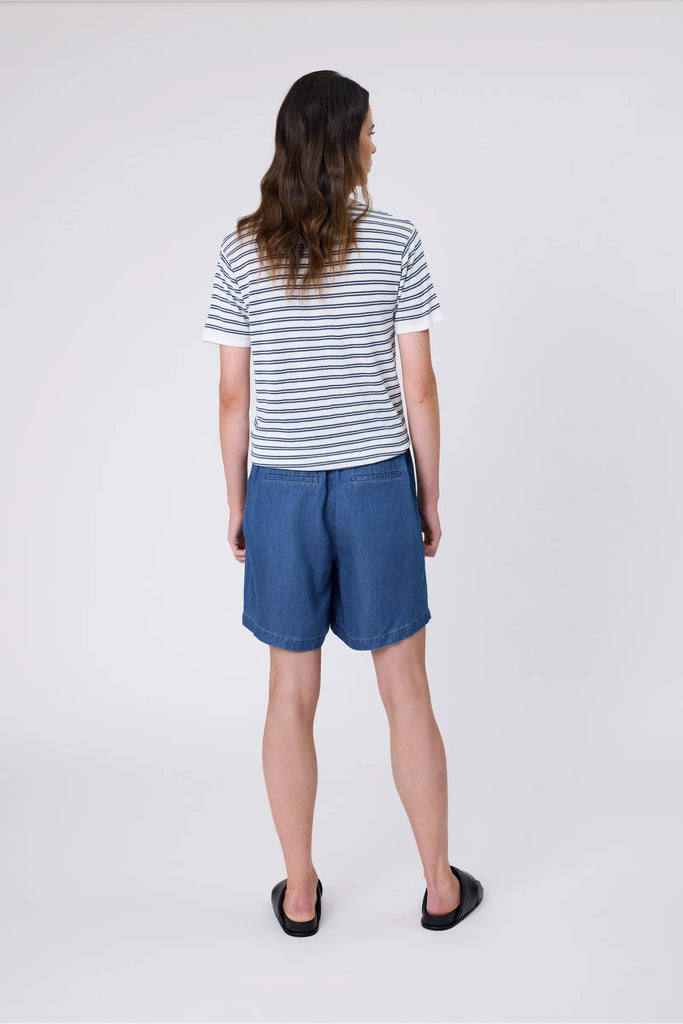 Woman wearing a striped polo shirt and blue shorts on a plain background. Marlow Chime Knit Polo Shirt in indigo Stripe.