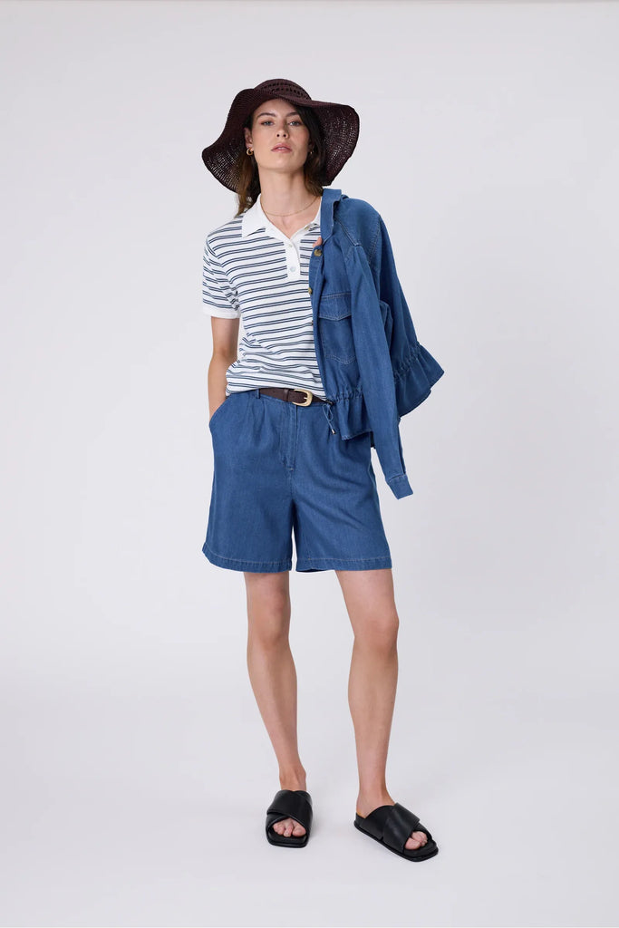 Woman wearing a striped polo shirt and blue shorts on a plain background. Marlow Chime Knit Polo Shirt in indigo Stripe.