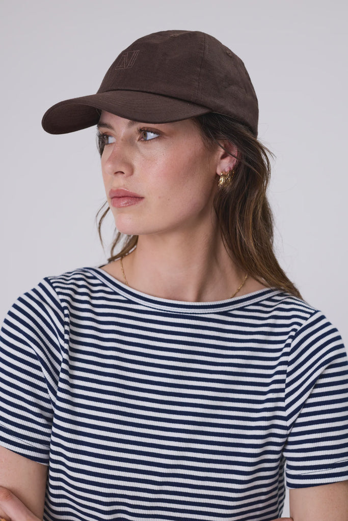 Woman wearing a brown cap and striped shirt against a plain background. Marlow Icon Linen Cap in carob.