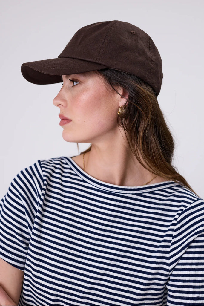 Woman wearing a brown cap and striped shirt against a plain background. Marlow Icon Linen Cap in carob.