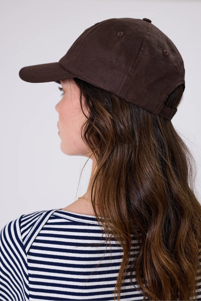 Woman wearing a brown cap and striped shirt against a plain background. Marlow Icon Linen Cap in carob.