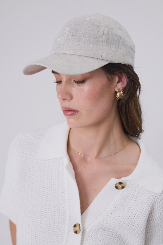 Woman wearing a light beige baseball cap and a white cardigan on a plain background. Marlow Icon Linen Cap Natural.