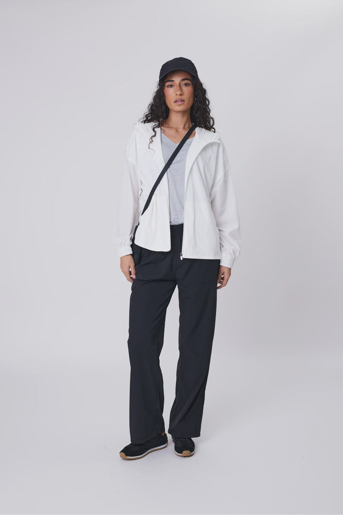 Woman wearing a white jacket, gray shirt, black pants, and black cap on a white background. Marlow Intrepid Pant 2.0 in Black
