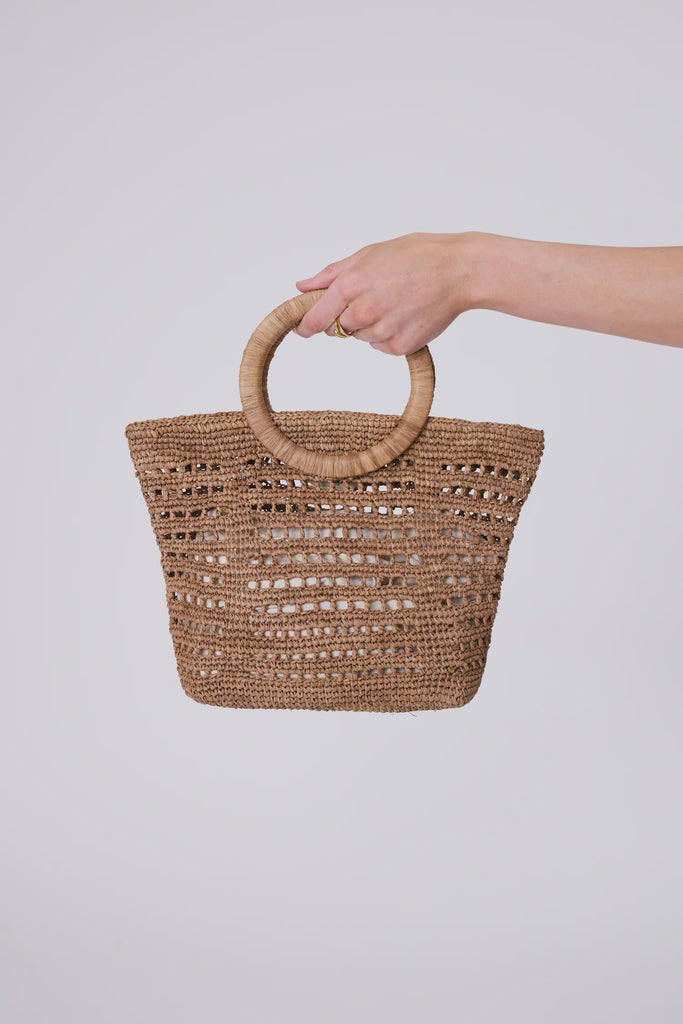 Woven raffia handbag  held by a hand on a light grey background. Milano Mini Bucket Bag in Tea.