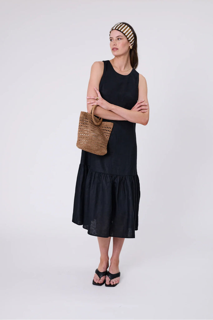 Woman holding a woven raffia handbag  wearing a black dress on a light grey background. Milano Mini Bucket Bag in Tea.