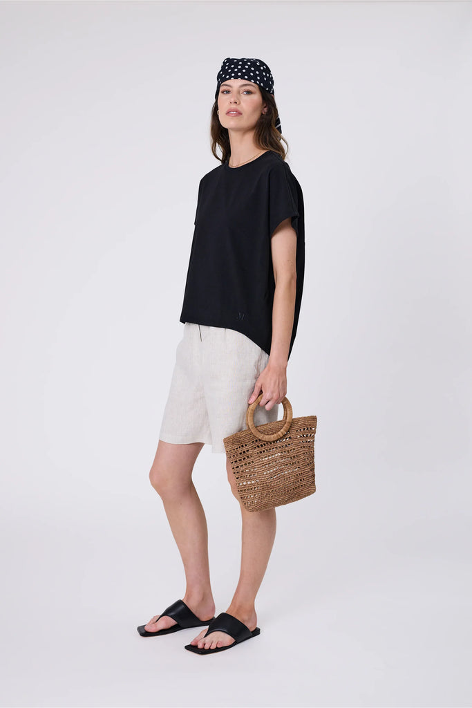 Woman holding a woven raffia handbag  wearing a black top on a light grey background. Milano Mini Bucket Bag in Tea.