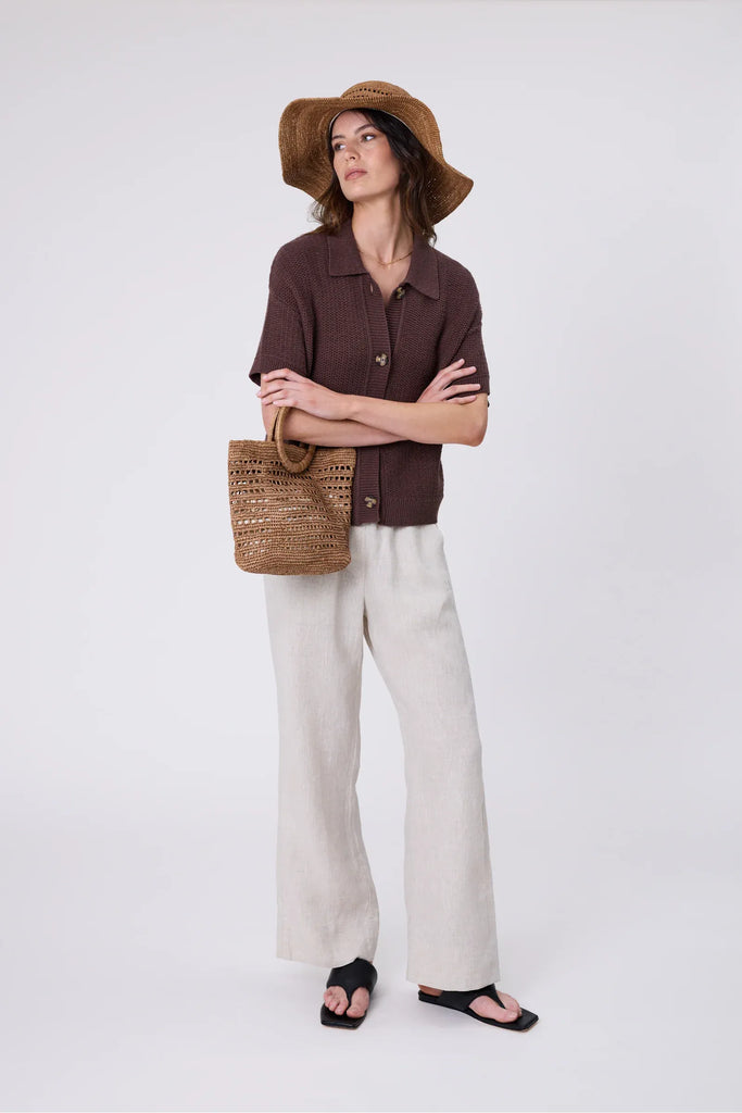 Woman holding a woven raffia handbag  wearing a brown top on a light grey background. Milano Mini Bucket Bag in Tea.