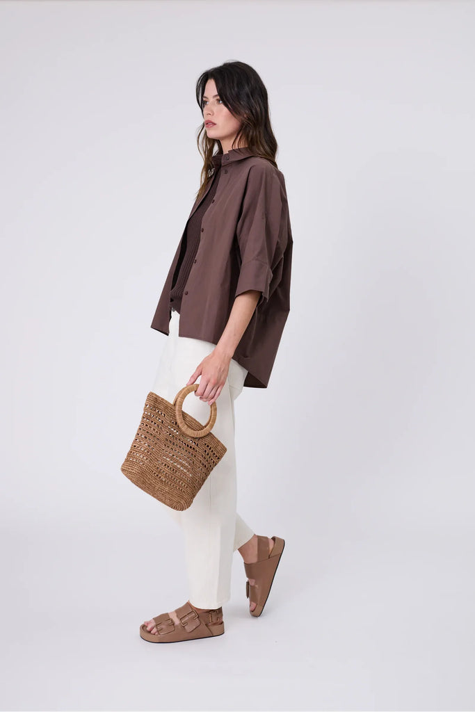 Woman holding a woven raffia handbag  wearing a brown shirt on a light grey background. Milano Mini Bucket Bag in Tea.