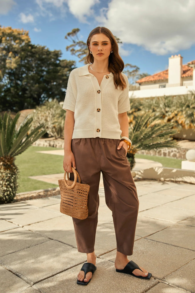 Woman holding a woven raffia handbag  wearing a beige dress on an outdoor background. Milano Mini Bucket Bag in Tea.