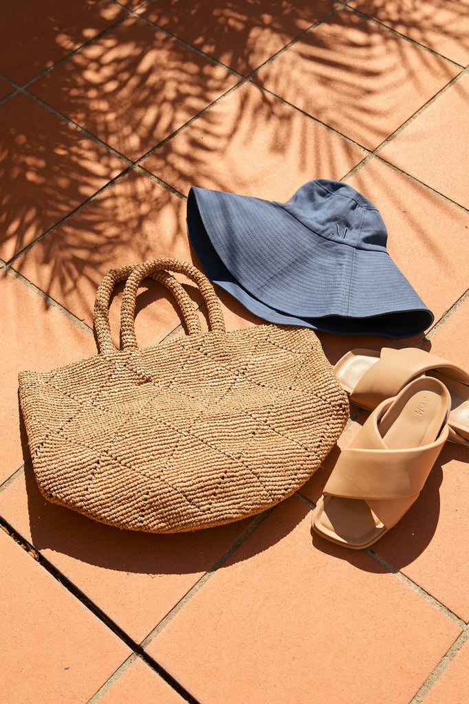 woven brown bag against a tiled background. Marlow Santorini Bucket Bag in tea.
