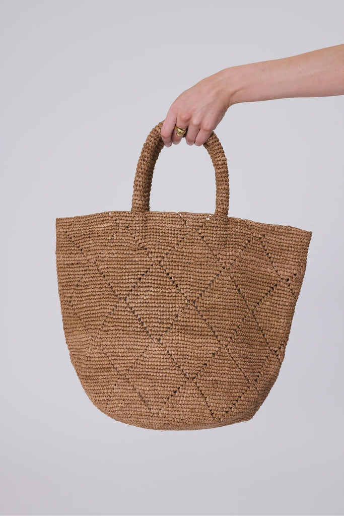 Hand holding a woven brown bag against a plain background. Marlow Santorini Bucket Bag in tea.