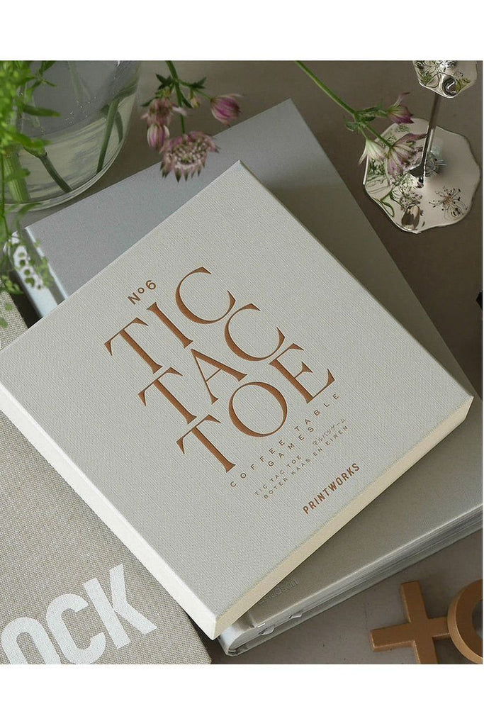 Printworks 'Tic Tac Toe' is presented in a book-like box sitting on a surface with flowers and a glass in the background.