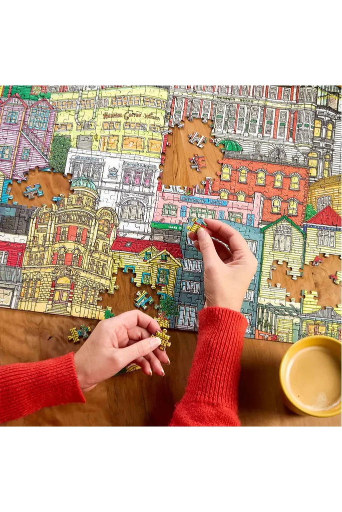 Piece House collaborates with Stacey Fraser Allen on a puzzle titled "Neighbourhood Puzzle." The image shows a hand assembling a detailed puzzle of a colourful cityscape on a wooden table.