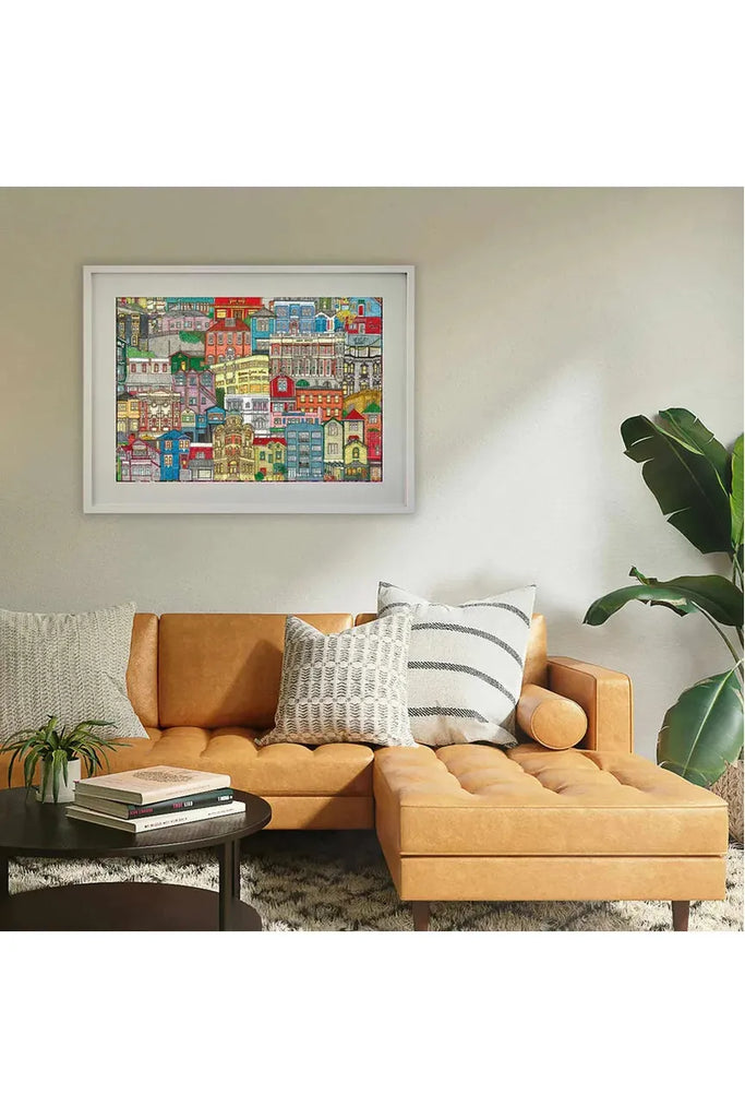 Piece House has teamed up with Stacey Fraser Allen to design a puzzle called "Neighbourhood Puzzle." The image features a lively cityscape of Wellington, with the finished puzzle beautifully framed and hung on a wall above a mustard yellow sectional sofa. The scene also includes decorative cushions and a small table with books.