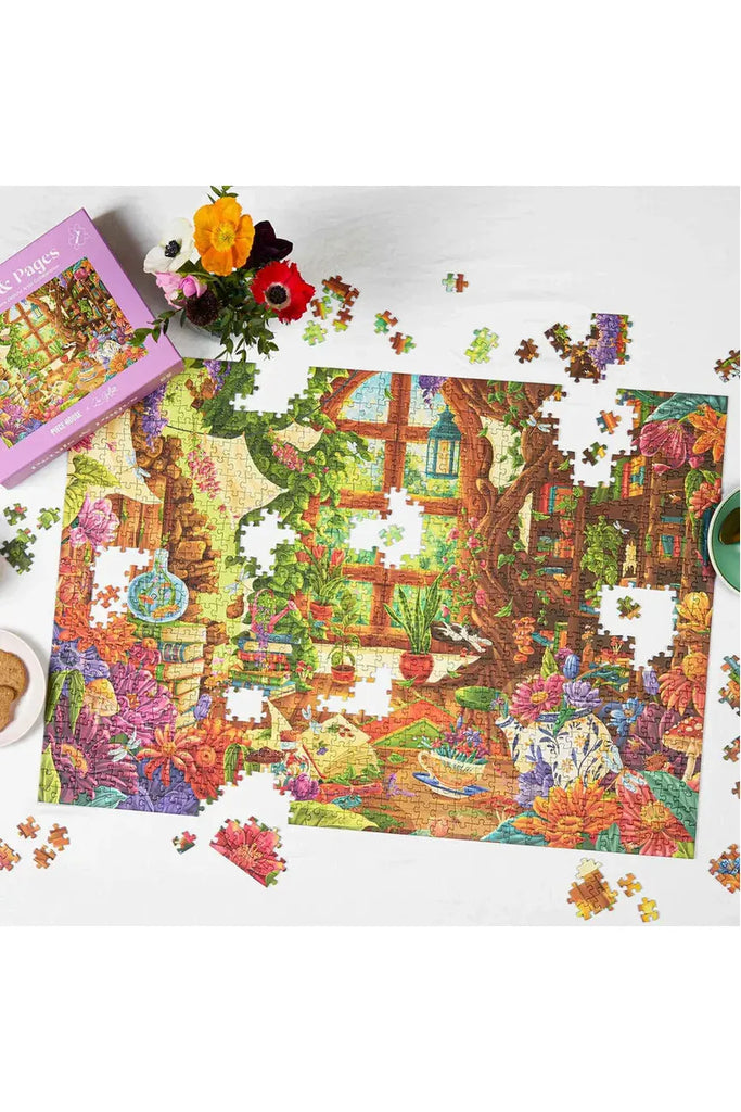 Piece House and Zoe Gillett's collaboration jigsaw puzzle titled 'Petals & Pages' features a cosy scene with books and plants arranged on a white surface, accompanied by puzzle pieces and a box.