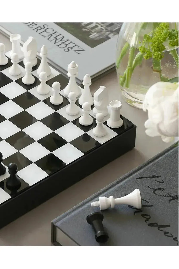 The Printworks Art of Chess set showcases elegant white pieces on a striking black and white checkered board, beautifully complemented by a charming vase of flowers in the background.