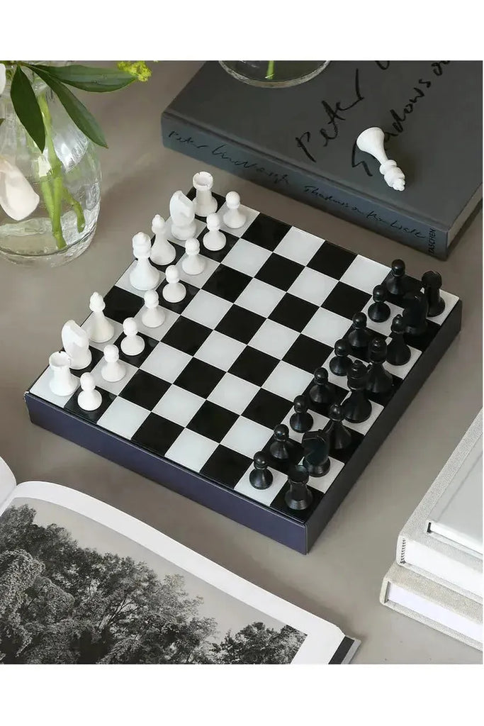Printworks Art of Chess, Chessboard with black and white pieces on a grey surface with books and a plant.