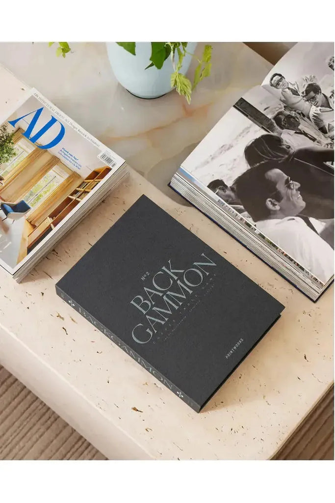 The Printworks Backgammon Classic Game Board resembles a book, featuring a dark grey background with turquoise text that displays the game’s name and the Printworks branding. "The Backgammon" is showcased on a marble surface along with magazines and a plant.