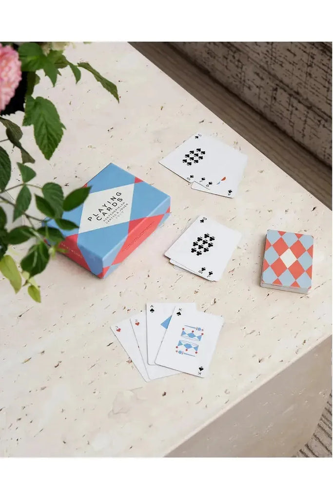Printworks Playing Cards are seen on a travertine table next to a plant. The cards feature a geometric design and are presented alongside their box.