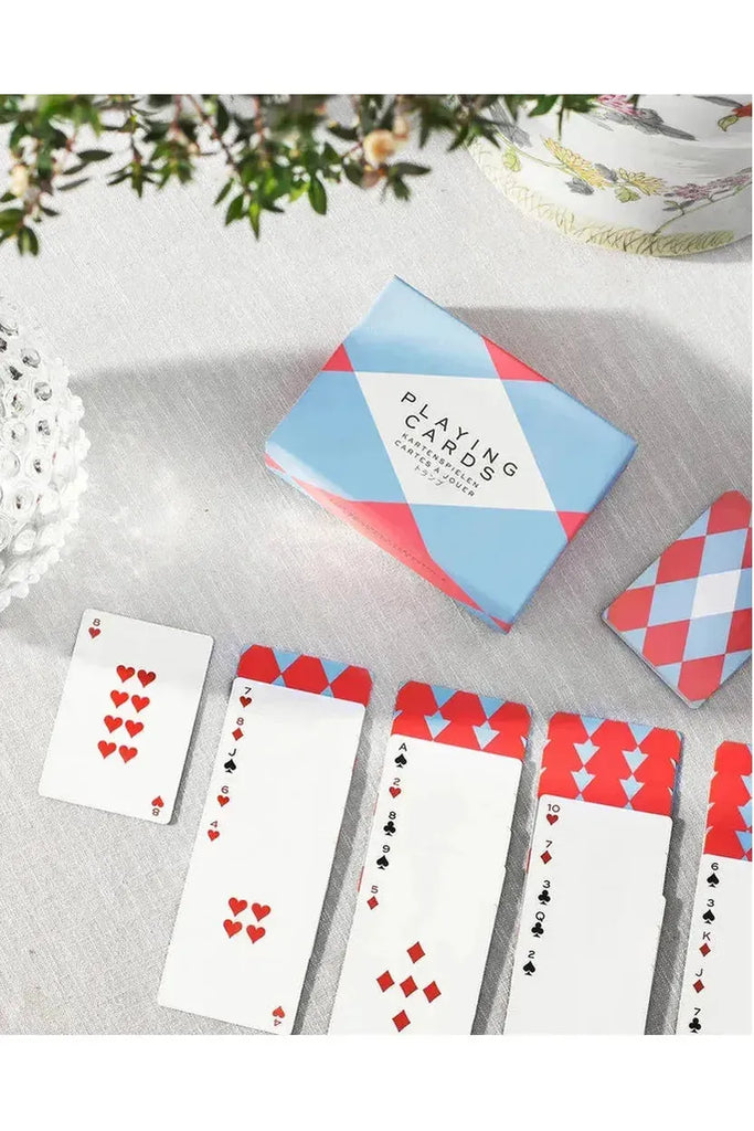 Printworks playing cards with a red, blue, and white design are laid out on a light surface, where someone is playing Patience. The presentation box features the same pattern alongside some greenery.