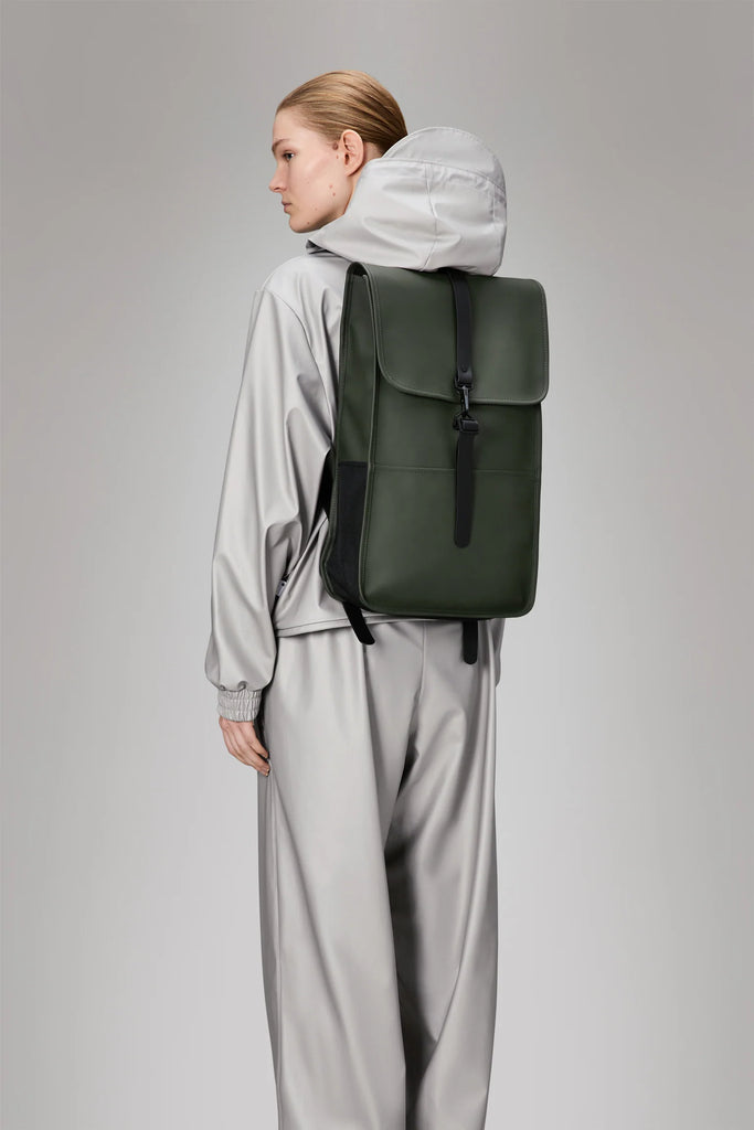Person wearing a khaki green backpack against a plain background. Rains Green backpack.