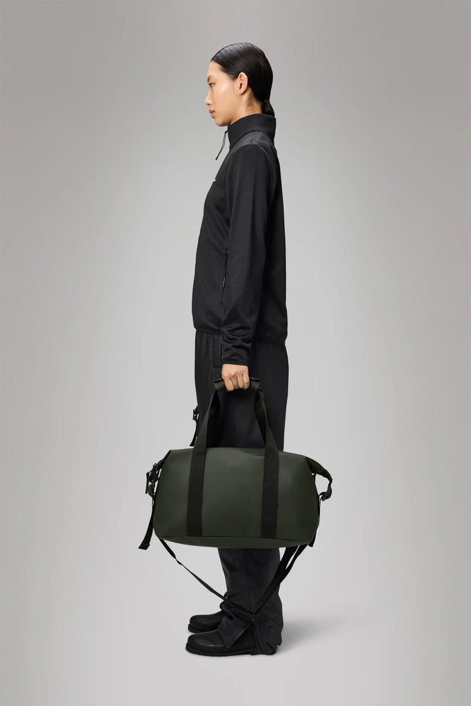 Person holding a Rains Green duffel weekend bag with black straps on a light gray background