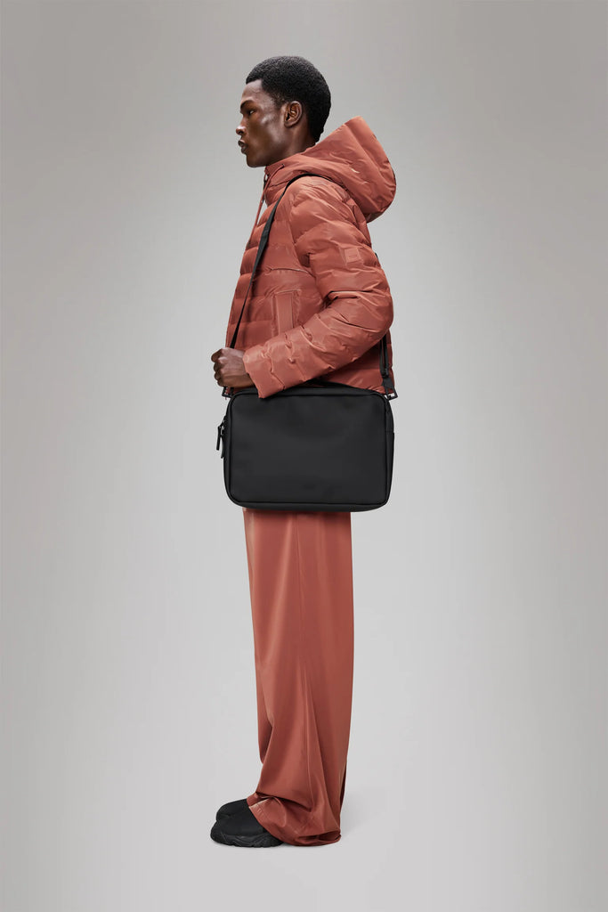 Person wearing a raincoat holding a Rains black laptop bag on a grey background