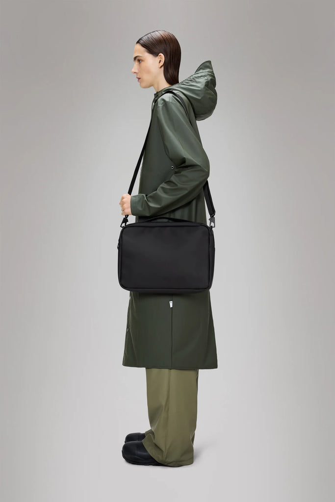 Person wearing a green raincoat holding a Rains black laptop bag on a grey background