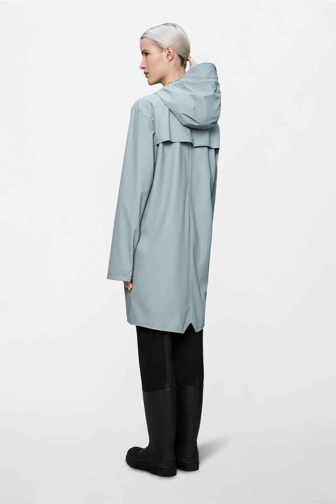 Person wearing a pale blue raincoat and holding a matching backpack on a light grey background. Rains Long Jacket in Pool.