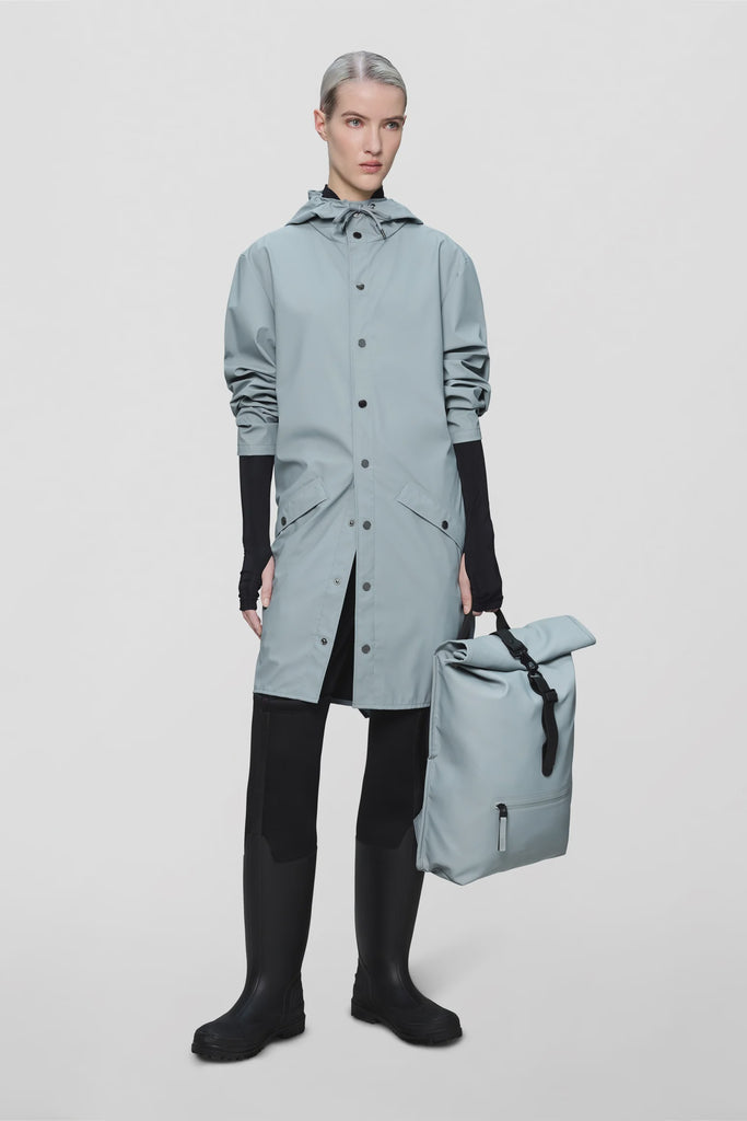 Person wearing a pale blue raincoat and holding a matching backpack on a light grey background. Rains Long Jacket in Pool.