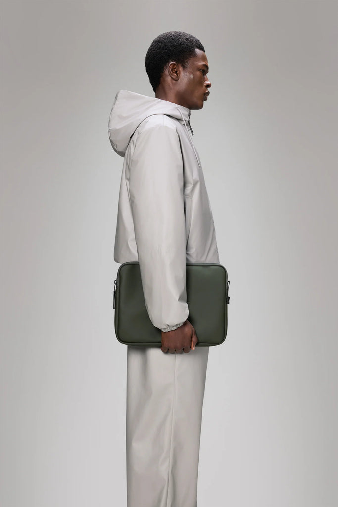 Person wearing a light gray jacket holding a Khaki green laptop case on a light grey background. Rains Trail Laptop Case in Green.